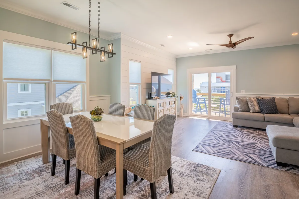 This interior shot encompasses a spacious dining area adjacent to a plush sofa, large windows, and a TV. The setup invites communal meals, festive gatherings, and seamless transitions to outdoor spaces, with sunlight pouring in to highlight the space's open-plan layout.