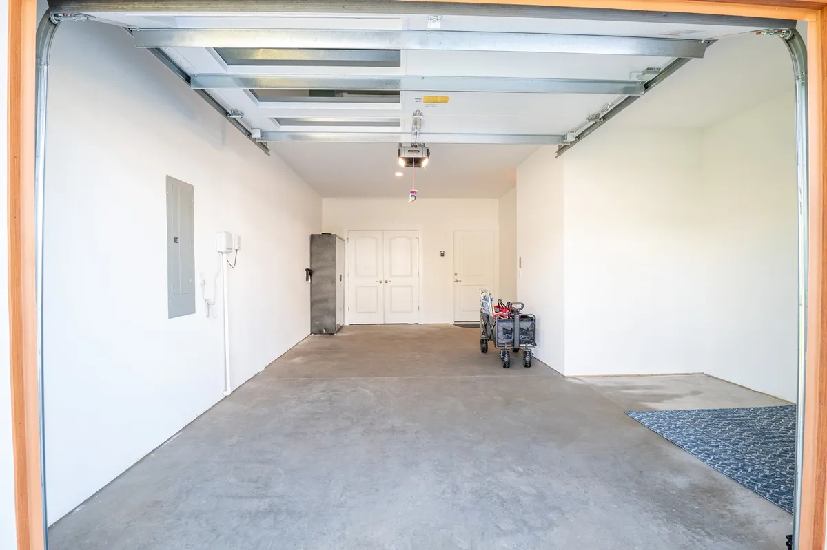 The image displays a spacious garage interior with a visible garage door, a utility panel, a refrigerator, and a door leading into the house. A storage cart adds a practical touch, suggesting ample storage and organization. The space feels uncluttered and ready for multiple vehicles or family gear.