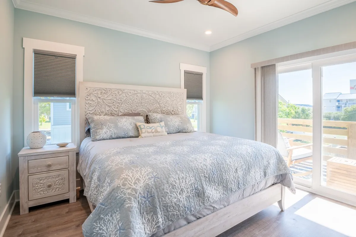A sunlit bedroom featuring a bed with an intricate headboard, plush decorative pillows, and large windows that fill the space with serene natural light. The room exudes calm and comfort, perfect for rest and relaxation.