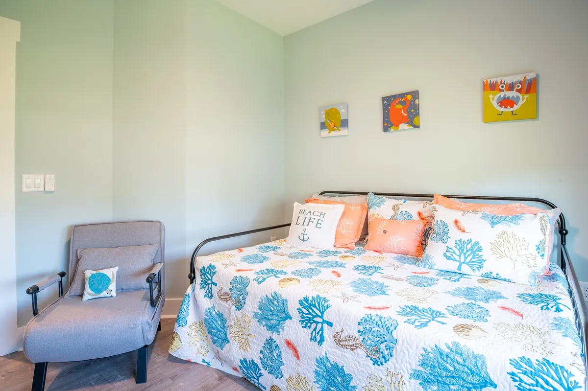 A playful, inviting kids' room featuring a daybed with a coral-patterned quilt, beach-themed pillows, colorful artwork, and a comfortable chair. The room is bright and cheerful, perfect for young travelers to relax, read, or play.