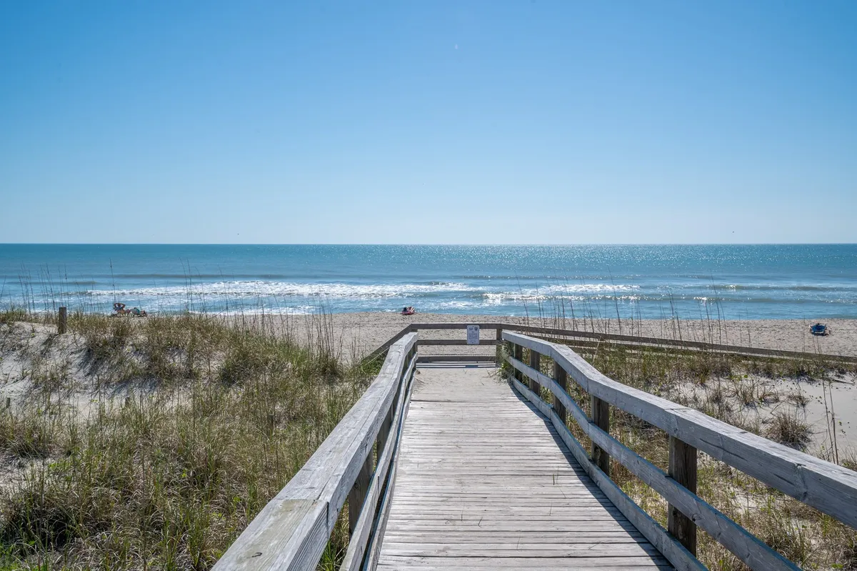 A sweeping exterior shot features a wide wooden boardwalk leading over dunes to a sandy beach, with gentle ocean waves rolling onto the shore and tufts of dune grass framing the scene. The area is sunlit, evoking an inviting, classic beach day atmosphere perfect for families.