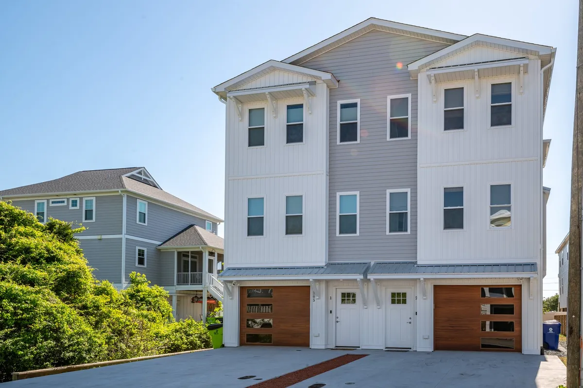 The image showcases the modern exterior of a newly built single-family house featuring sleek architectural lines, warm wooden garage doors, expansive windows, and an inviting porch perfect for gatherings. Sunlight highlights the contemporary design, while the porch hints at relaxation and socializing, all set just three blocks from the Carolina Beach shoreline.