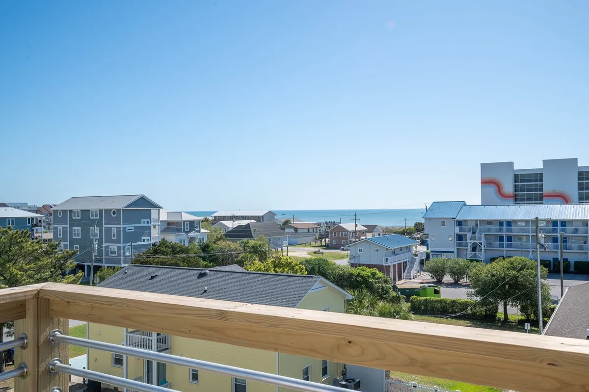 A serene balcony view shows the ocean in the distance, colorful beach houses, and a peaceful neighborhood landscape. The scene captures a quiet morning or evening, hinting at relaxing moments spent watching the sunrise or sunset over the water.