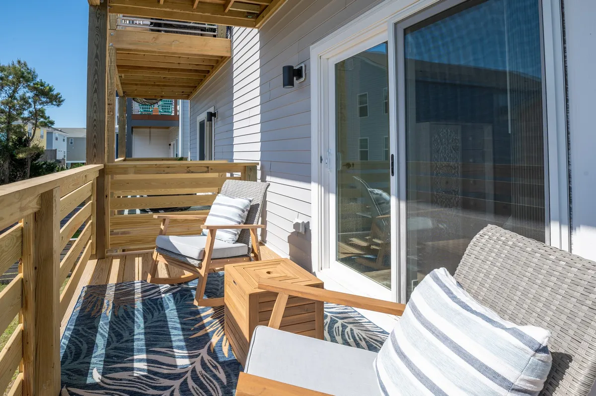 A serene exterior scene featuring a wooden railing, outdoor chairs, a small table, and a rug on a balcony or deck. The setup invites guests to relax while enjoying the fresh air and view, with sunlight suggesting a pleasant outdoor atmosphere.