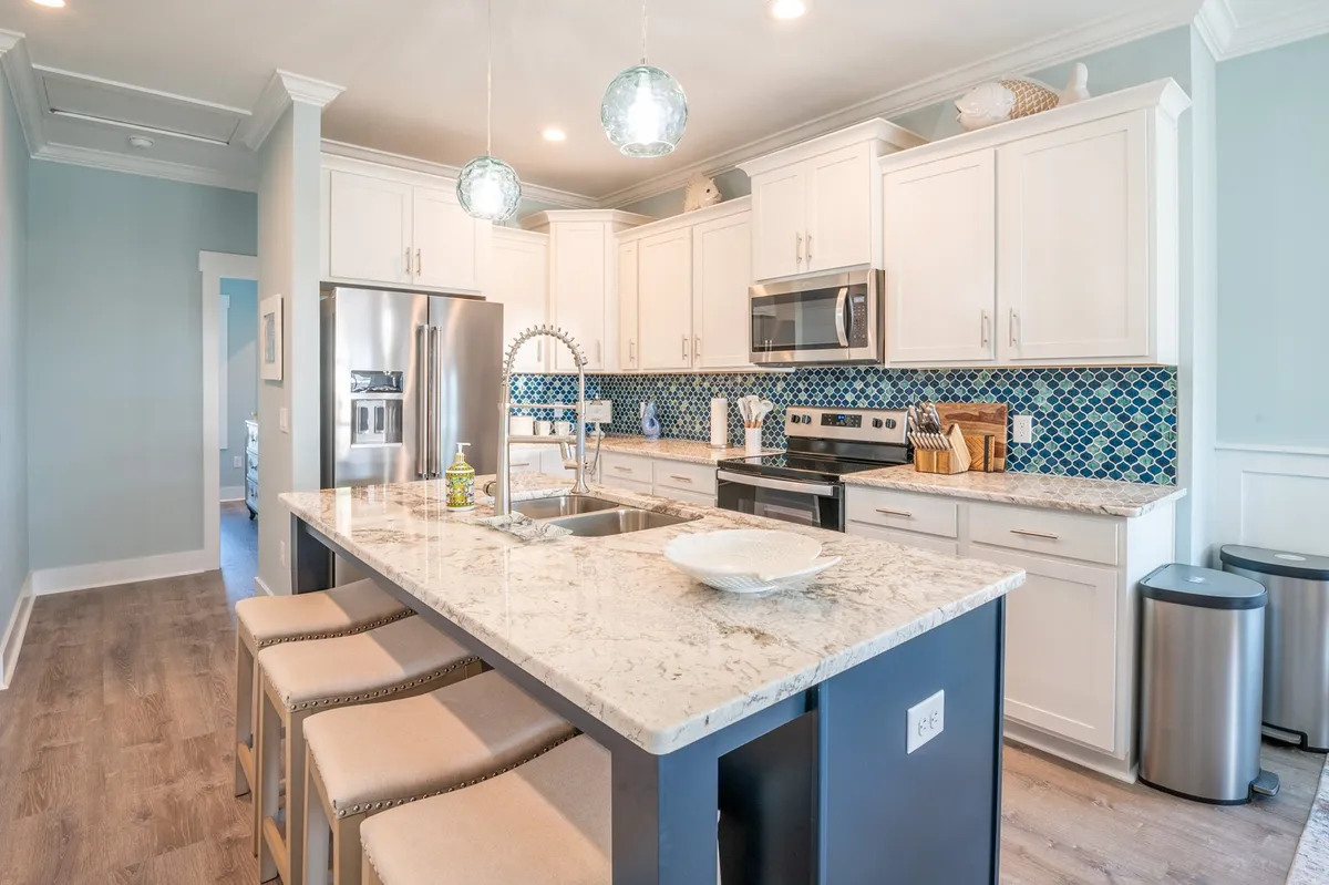 A modern kitchen with a granite countertop, blue backsplash, island seating, and pendant lights. The room is well-lit and designed for cooking, entertaining, and gathering with friends or family.