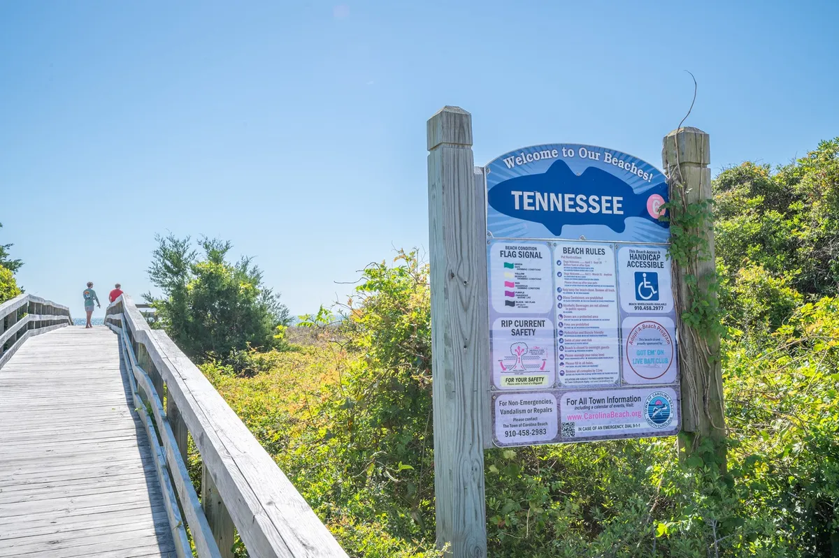 A welcoming shot of the Carolina Beach entrance features a prominent 'Welcome' sign, a classic wooden boardwalk, and people strolling toward the ocean. The environment feels lively yet relaxed, capturing the friendly, communal atmosphere of the area.