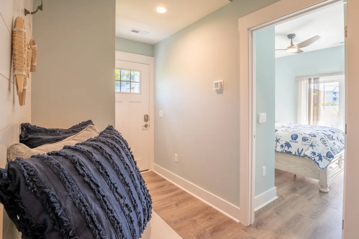 This image features a cozy and inviting entryway decorated with blue textured pillows, an entrance door, and a passage leading to a bedroom. The scene conveys the sense of coming home after a day at the beach, with soft lighting and a welcoming ambiance.