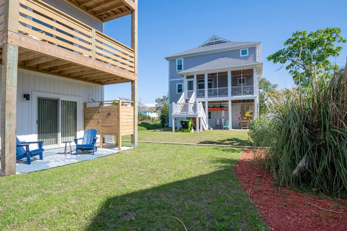 An exterior view showcasing a deck area with comfortable seating, a large house in the background, and lush green grass. The mood is cheerful and relaxed, ideal for returning from the beach and unwinding outdoors.